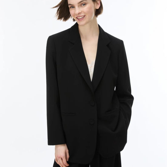 A woman modeling a black tailored blazer with wide lapels, two front buttons, and large patch pockets, paired with black trousers against a clean white studio background.