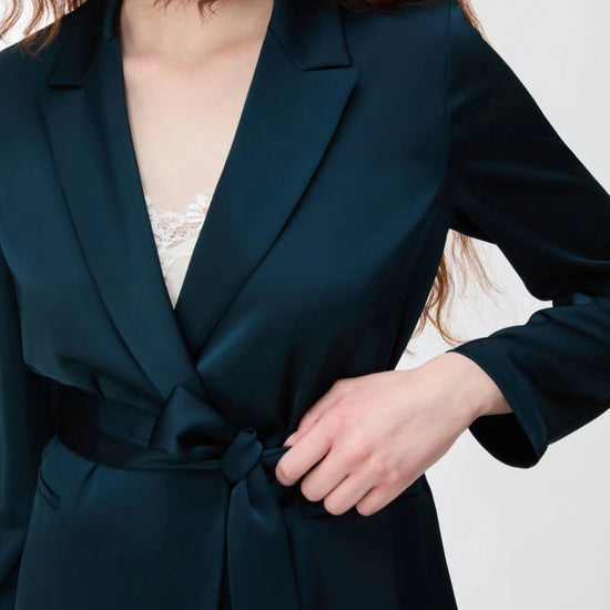 A close-up of a model wearing a dark teal satin wrap jacket with a tie waist belt, styled over a white lace camisole against a light gray background.