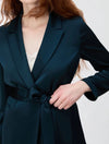A close-up of a model wearing a dark teal satin wrap jacket with a tie waist belt, styled over a white lace camisole against a light gray background.
