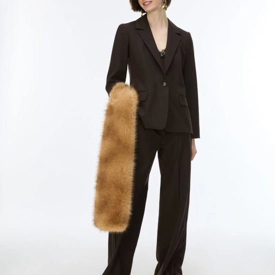 A model wearing a dark brown tailored suit with wide-leg trousers and sunglasses, holding a long, plush tan fur accessory against a plain white studio background.