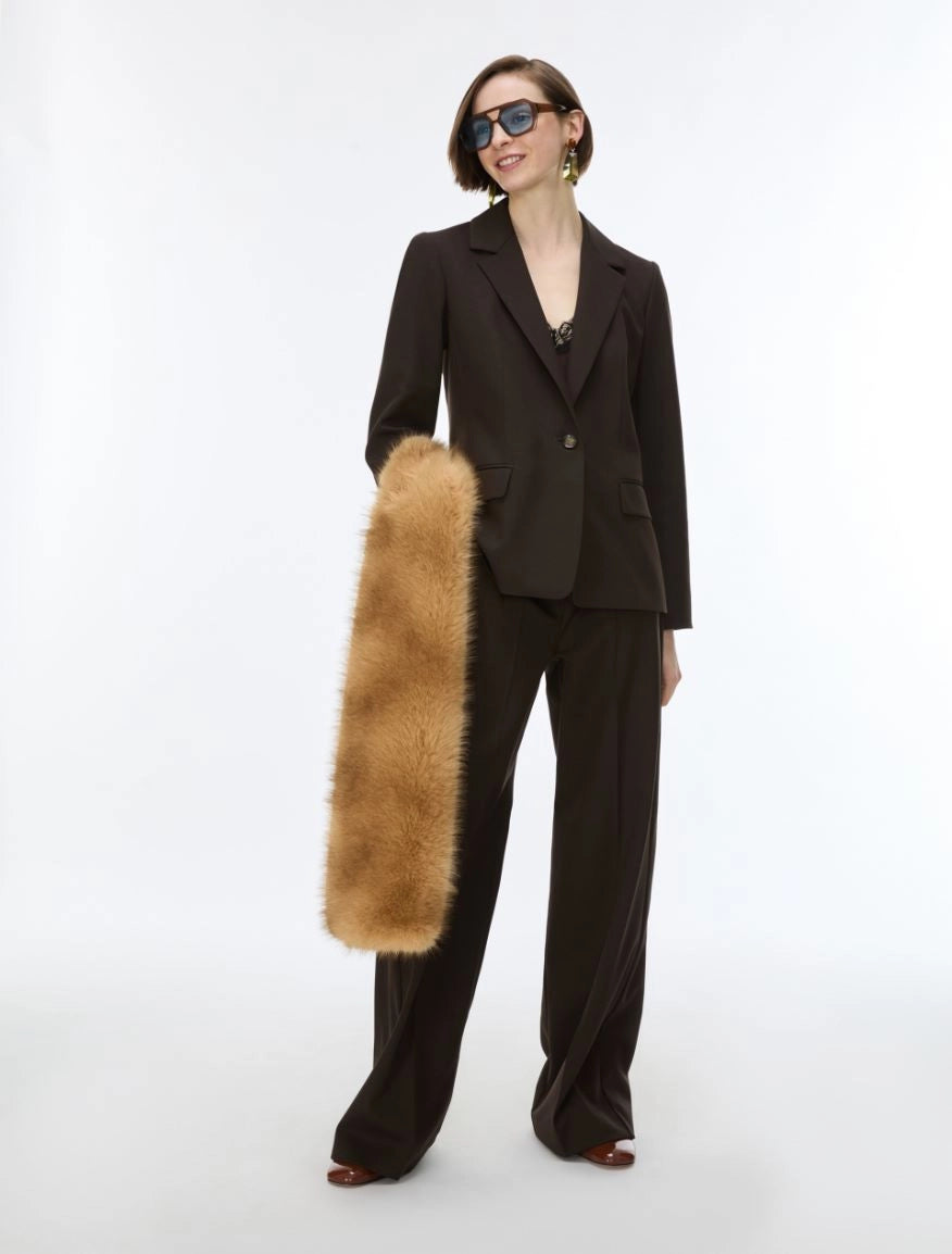 A model wearing a dark brown tailored suit with wide-leg trousers and sunglasses, holding a long, plush tan fur accessory against a plain white studio background.