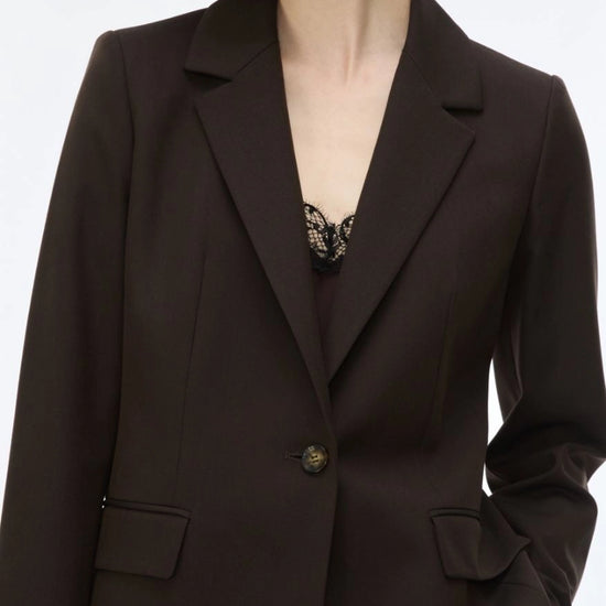 A close-up of a model wearing a dark brown tailored blazer with notch lapels, flap pockets, and a single button closure over a black lace top.