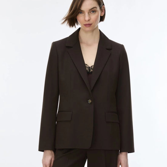 A woman modeling a dark brown tailored blazer with matching trousers against a plain light gray studio background.