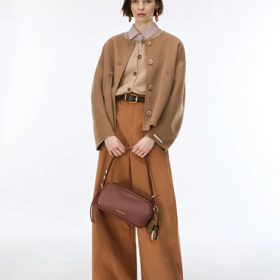 A model stands against a light gray studio background wearing a camel cropped jacket over a beige collared shirt, wide-leg brown trousers, and a brown crossbody bag.