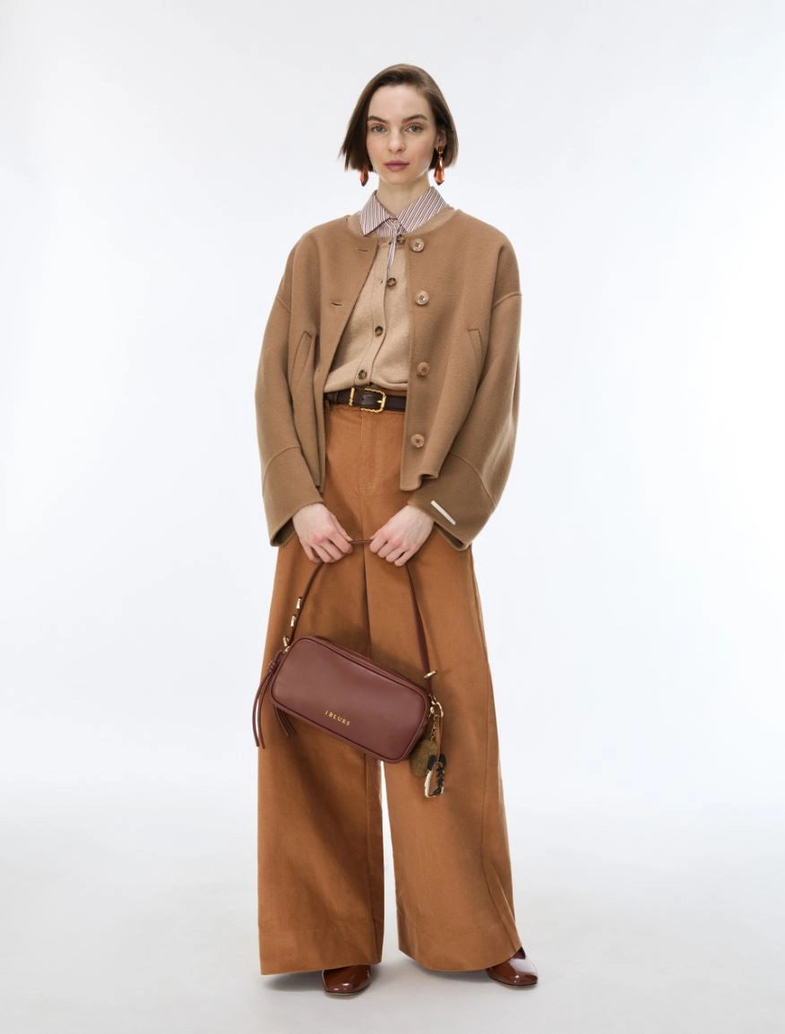A model stands against a light gray studio background wearing a camel cropped jacket over a beige collared shirt, wide-leg brown trousers, and a brown crossbody bag.