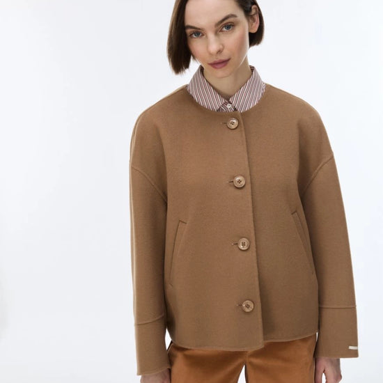 A woman modeling a camel brown cropped button-up cardigan over a striped collared shirt and brown trousers, shot against a plain white studio background.