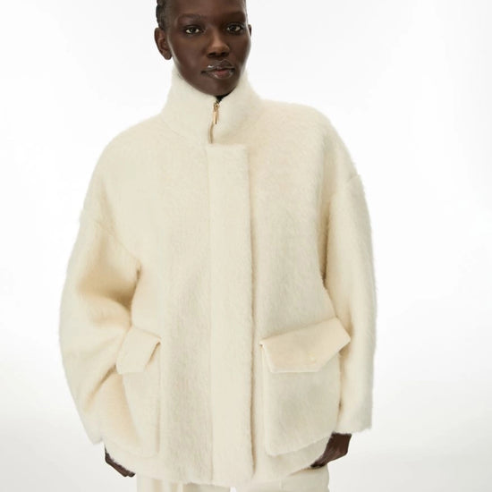 A model wearing a cream-colored, fuzzy cropped jacket with a high stand-up collar and large front patch pockets, paired with matching cream trousers, against a plain white studio background.