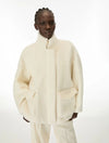 A model wearing a cream-colored, fuzzy cropped jacket with a high stand-up collar and large front patch pockets, paired with matching cream trousers, against a plain white studio background.
