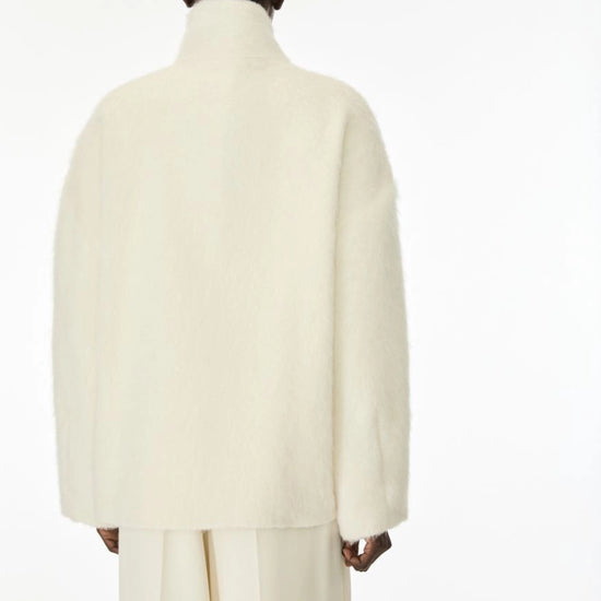 A model shown from the back wearing a voluminous, high-neck cream-colored coat and matching trousers against a plain white studio background.