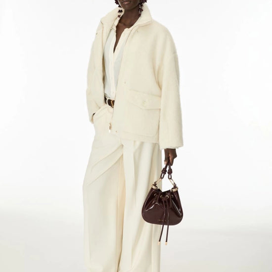A model stands against a plain white studio background wearing a cream-colored two-piece jacket and wide-leg trousers set, holding a dark brown leather bucket bag.