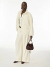 A model stands against a plain white studio background wearing a cream-colored two-piece jacket and wide-leg trousers set, holding a dark brown leather bucket bag.