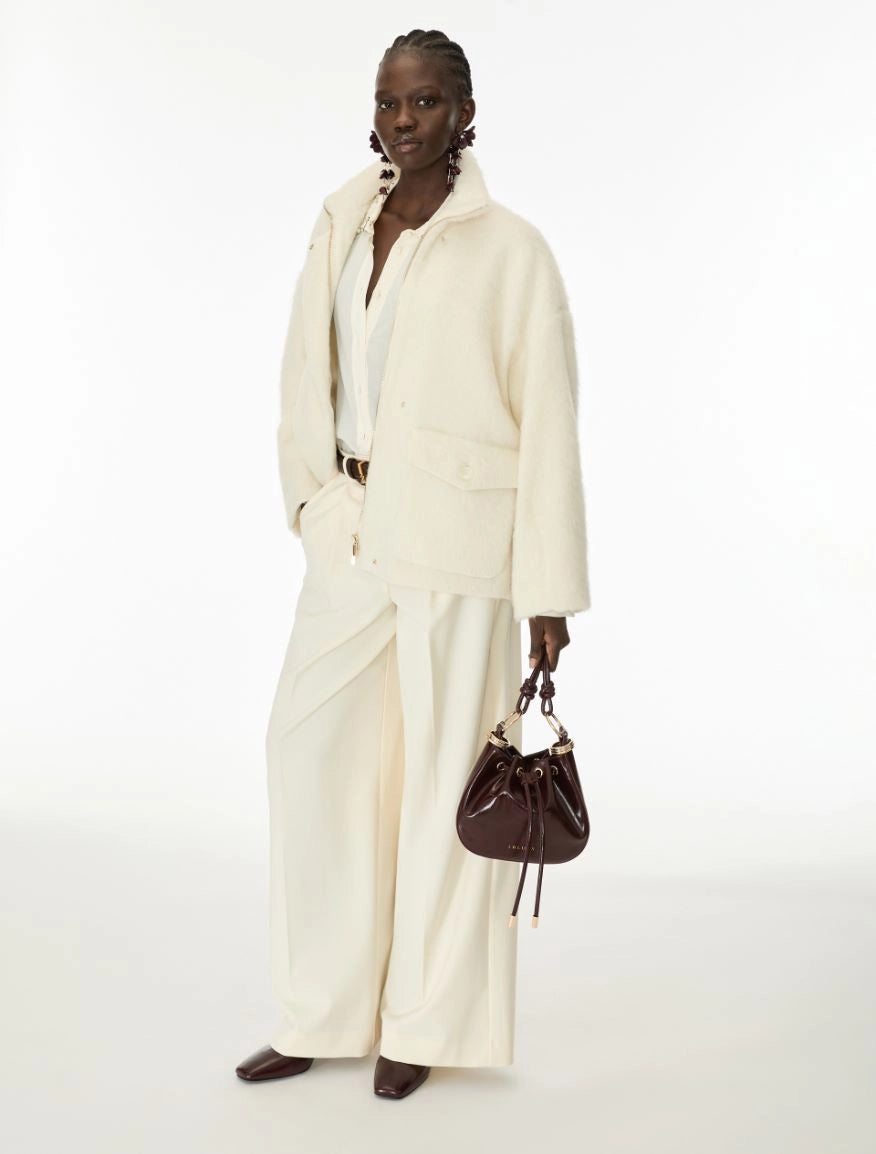 A model stands against a plain white studio background wearing a cream-colored two-piece jacket and wide-leg trousers set, holding a dark brown leather bucket bag.