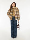 A woman models a stylish outfit featuring a beige and brown plaid fuzzy jacket, a tan knit sweater, dark blue patterned wide-leg trousers, and a small dark brown handbag, set against a plain white studio background.