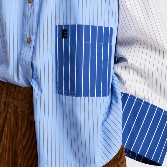 A model wearing a light blue and white color-blocked long-sleeve shirt with blue striped details, paired with wide-leg brown corduroy trousers, black sunglasses, and black shoes, holding a red and blue bag against a plain white studio background.