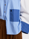 A model wearing a light blue and white color-blocked long-sleeve shirt with blue striped details, paired with wide-leg brown corduroy trousers, black sunglasses, and black shoes, holding a red and blue bag against a plain white studio background.