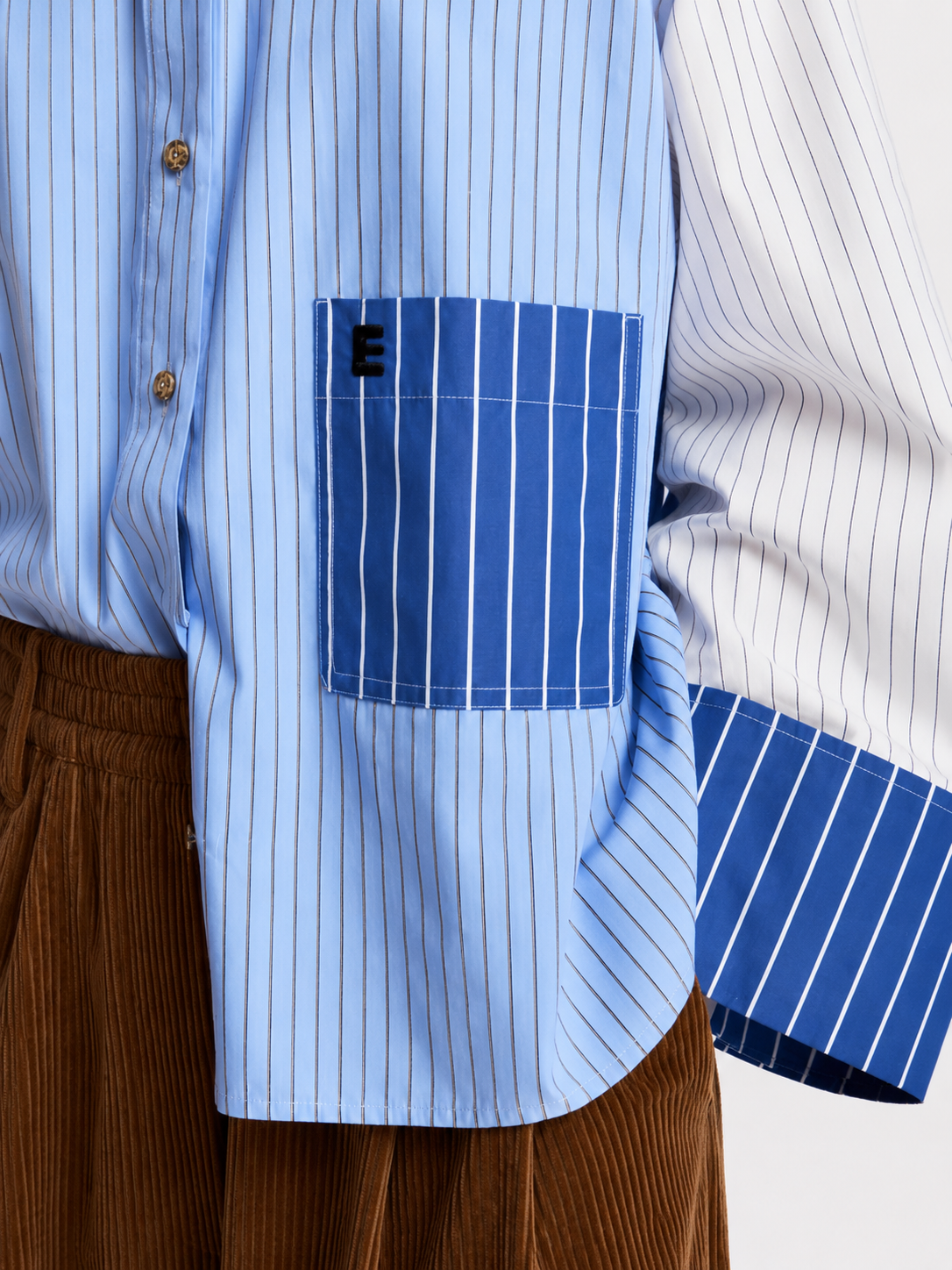A model wearing a light blue and white color-blocked long-sleeve shirt with blue striped details, paired with wide-leg brown corduroy trousers, black sunglasses, and black shoes, holding a red and blue bag against a plain white studio background.
