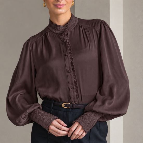 A woman wearing a dark brown long-sleeve blouse with a ruffled mock neck and button placket, paired with dark trousers and a belt, standing against a neutral gray background.