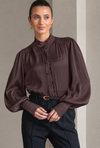 A woman wearing a dark brown long-sleeve blouse with a ruffled mock neck and button placket, paired with dark trousers and a belt, standing against a neutral gray background.
