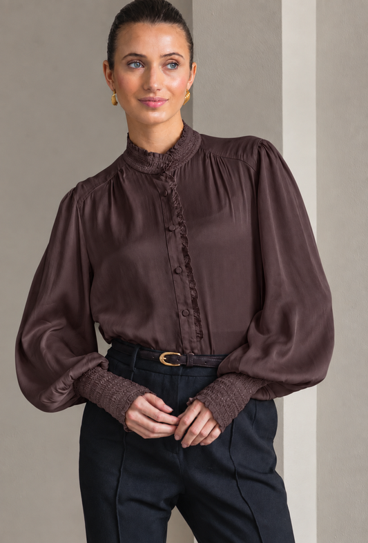 A woman wearing a dark brown long-sleeve blouse with a ruffled mock neck and button placket, paired with dark trousers and a belt, standing against a neutral gray background.