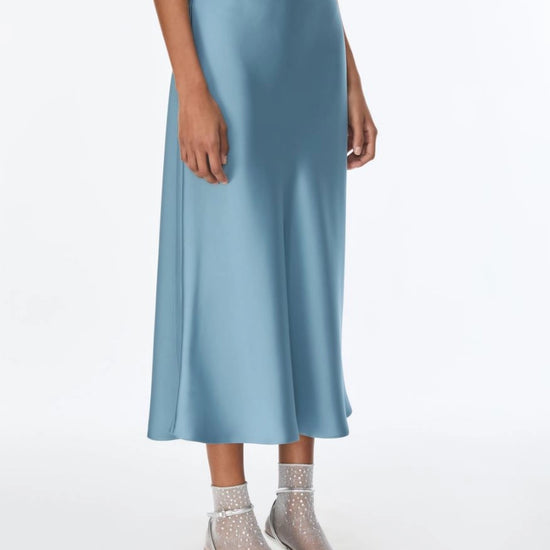 A model wearing a light blue satin midi skirt with a matching top and sheer high-heeled sandals, shown from the waist down on a white studio background.