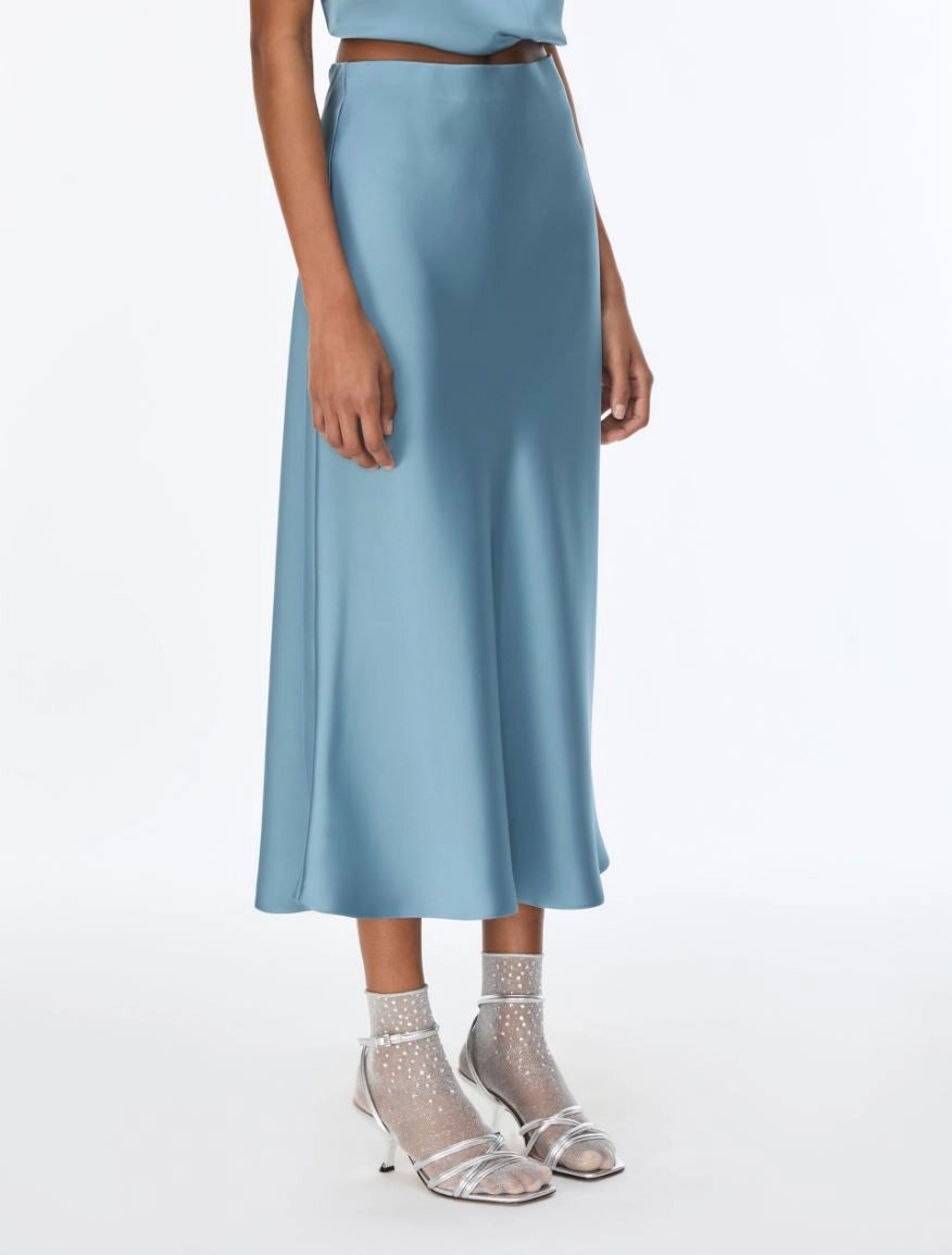 A model wearing a light blue satin midi skirt with a matching top and sheer high-heeled sandals, shown from the waist down on a white studio background.