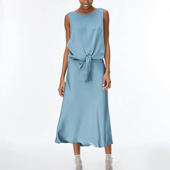 A woman modeling a sleeveless light blue midi dress with a knotted waist and tie detail, paired with clear high-heeled sandals, against a plain white studio background.