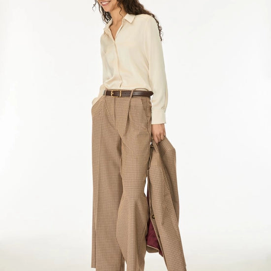 A woman modeling a cream long-sleeve button-up shirt, beige wide-leg plaid trousers, a brown belt, and holding a matching plaid coat, against a plain white studio background.