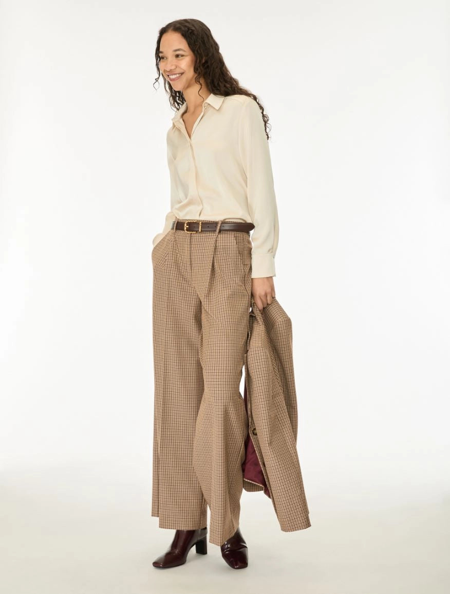 A woman modeling a cream long-sleeve button-up shirt, beige wide-leg plaid trousers, a brown belt, and holding a matching plaid coat, against a plain white studio background.