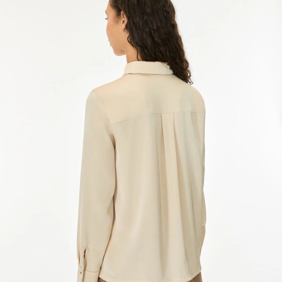 A woman shown from the back wearing a light beige long-sleeve blouse and brown checkered trousers against a plain white studio background.