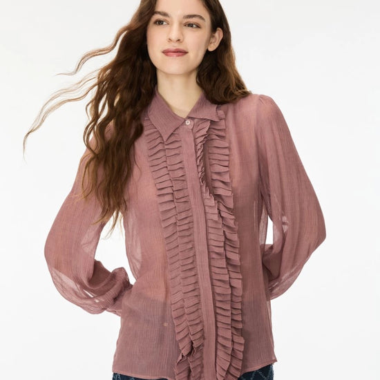 A woman modeling a dusty rose long-sleeve blouse with a ruffled front panel and collar, paired with dark blue patterned pants, against a plain white background.