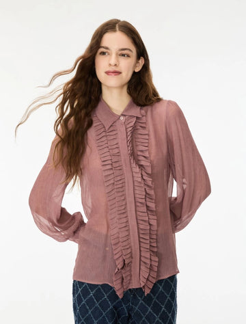 A woman modeling a dusty rose long-sleeve blouse with a ruffled front panel and collar, paired with dark blue patterned pants, against a plain white background.
