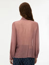 A woman viewed from behind wearing a sheer, long-sleeve dusty rose blouse and dark blue patterned trousers, set against a plain white studio background.