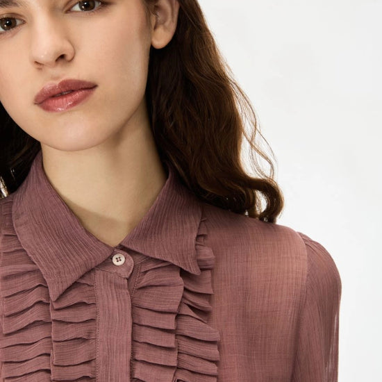 Close-up of a woman wearing a dusty rose ruffled blouse with a pointed collar and button placket, photographed against a clean white background.