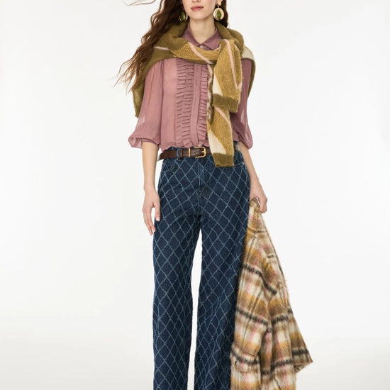 A woman models a coordinated outfit against a plain light background, wearing a dusty pink ruffled blouse, dark blue diamond-patterned wide-leg pants, a mustard green scarf, and holding a beige plaid shawl.