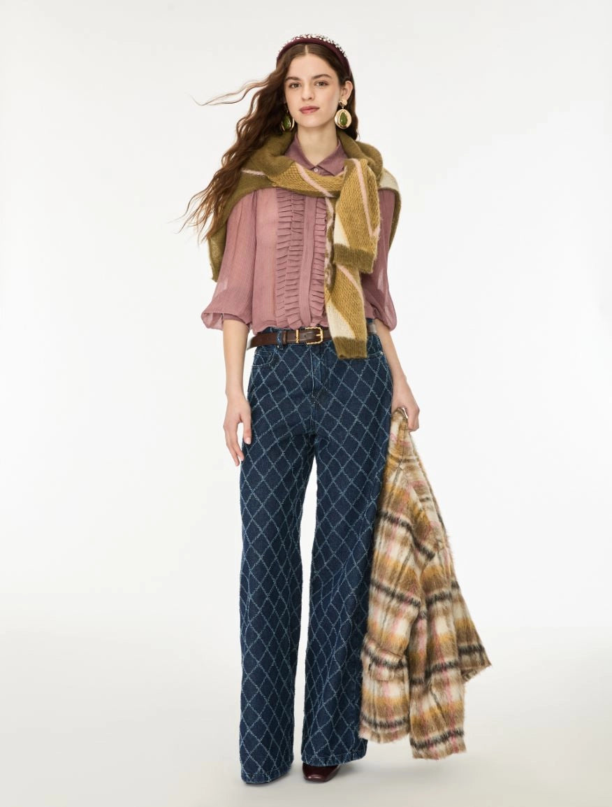 A woman models a coordinated outfit against a plain light background, wearing a dusty pink ruffled blouse, dark blue diamond-patterned wide-leg pants, a mustard green scarf, and holding a beige plaid shawl.