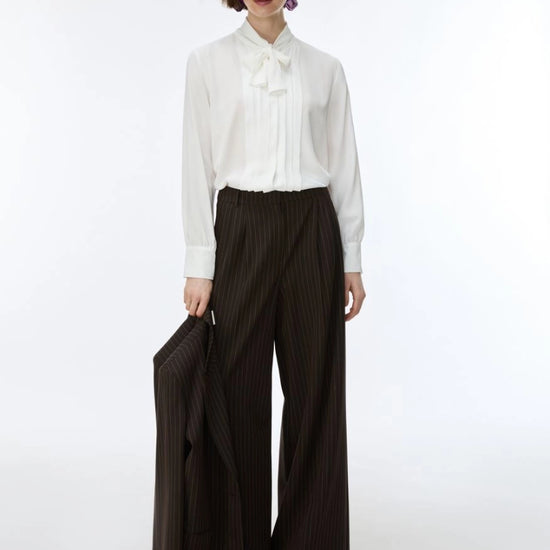 A model stands against a plain white studio background wearing a white long-sleeve blouse with a ruffled bow collar and dark brown wide-leg pinstriped trousers, holding a matching dark brown pinstriped blazer.