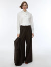 A model stands against a plain white studio background wearing a white long-sleeve blouse with a ruffled bow collar and dark brown wide-leg pinstriped trousers, holding a matching dark brown pinstriped blazer.