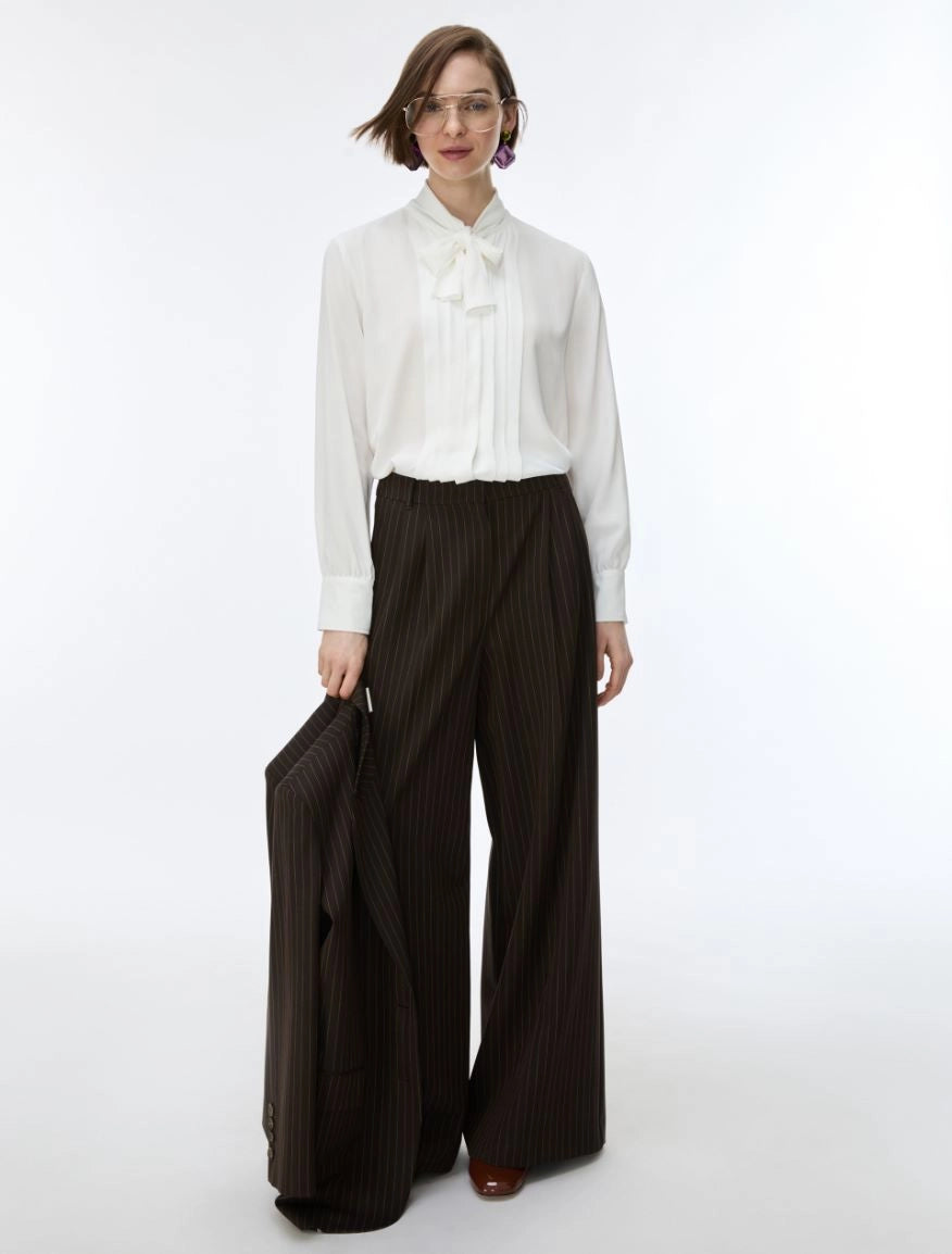 A model stands against a plain white studio background wearing a white long-sleeve blouse with a ruffled bow collar and dark brown wide-leg pinstriped trousers, holding a matching dark brown pinstriped blazer.