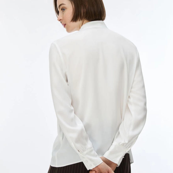 A woman with a short brown bob haircut is shown from the back wearing a white long-sleeve button-up shirt and dark brown pinstriped trousers, set against a plain white studio background.