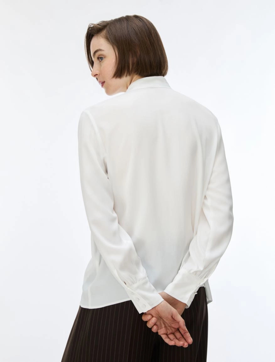 A woman with a short brown bob haircut is shown from the back wearing a white long-sleeve button-up shirt and dark brown pinstriped trousers, set against a plain white studio background.