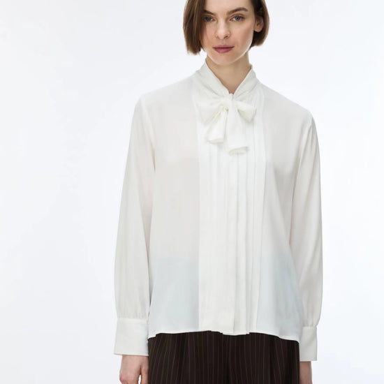 A woman wearing a white long-sleeve blouse with a high pleated collar and bow detail, paired with dark pleated trousers, against a plain white studio background.