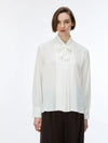 A woman wearing a white long-sleeve blouse with a high pleated collar and bow detail, paired with dark pleated trousers, against a plain white studio background.