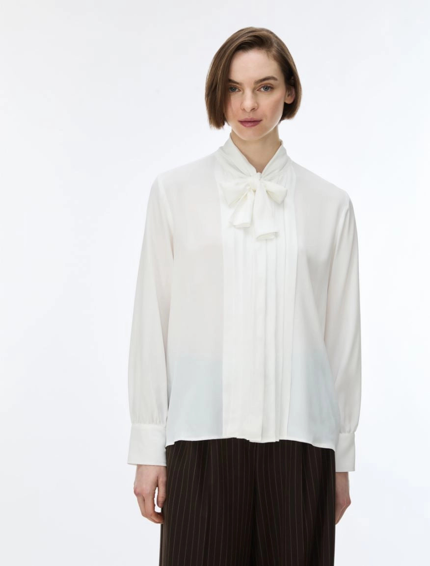 A woman wearing a white long-sleeve blouse with a high pleated collar and bow detail, paired with dark pleated trousers, against a plain white studio background.