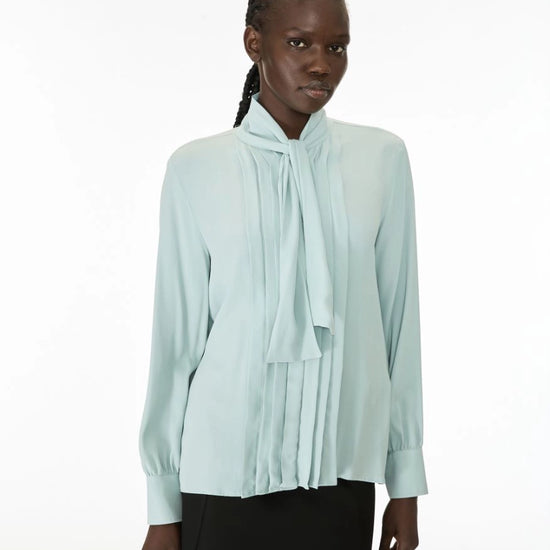 A model wearing a pale mint green long-sleeve blouse with a high gathered collar and a large front bow, paired with a black skirt, against a plain white studio background.