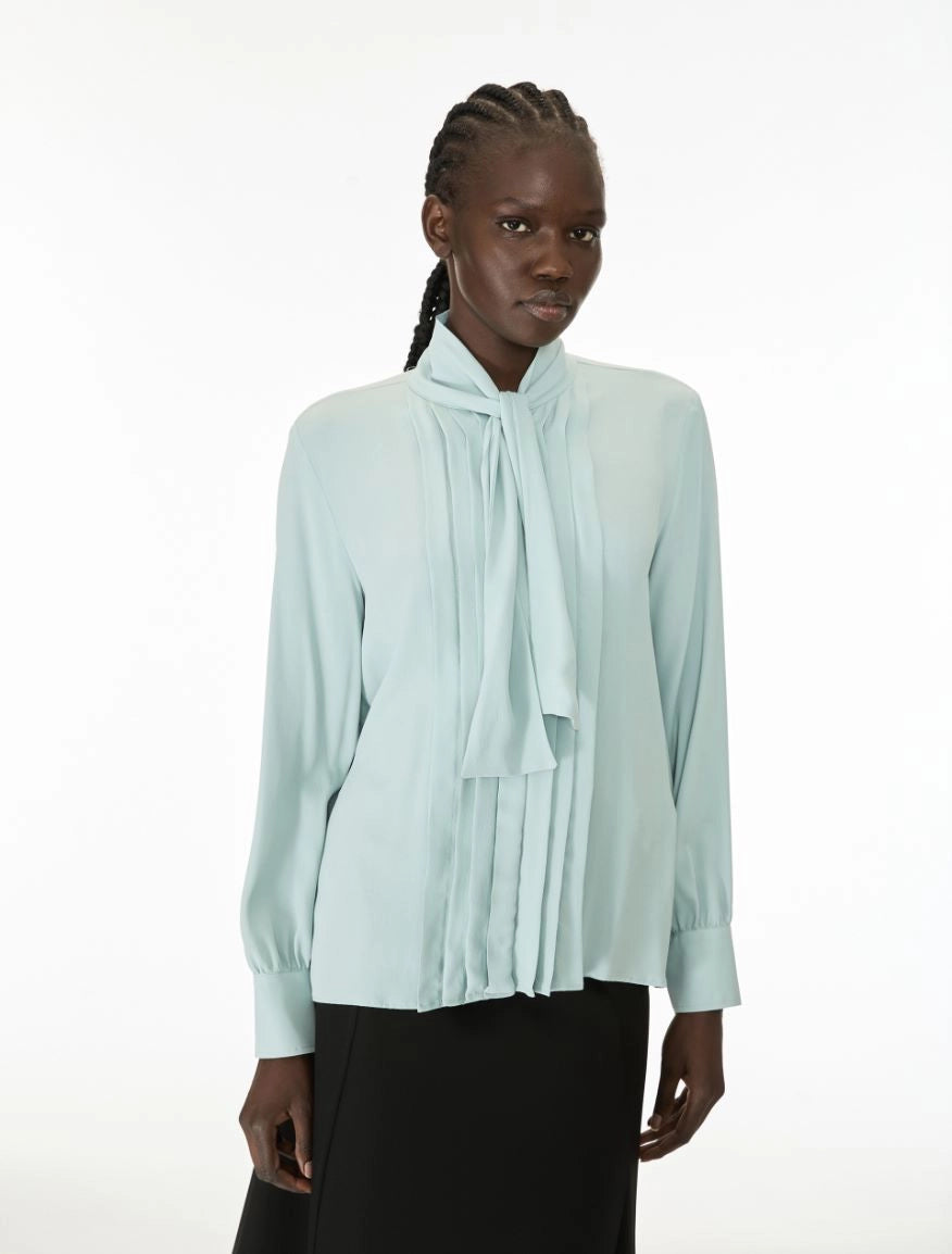 A model wearing a pale mint green long-sleeve blouse with a high gathered collar and a large front bow, paired with a black skirt, against a plain white studio background.