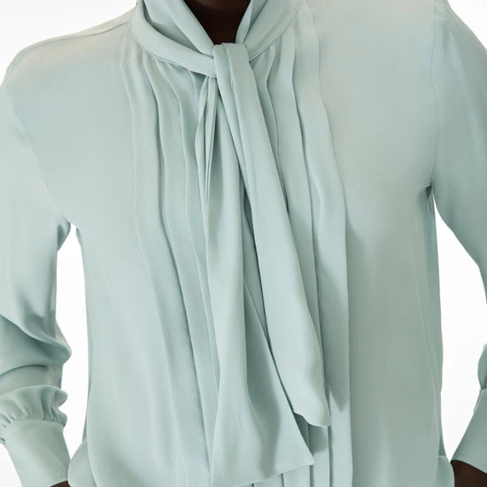 A close-up of a model wearing a pale mint green long-sleeve blouse with a high draped collar and a large front tie, set against a plain white background.