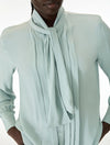 A close-up of a model wearing a pale mint green long-sleeve blouse with a high draped collar and a large front tie, set against a plain white background.