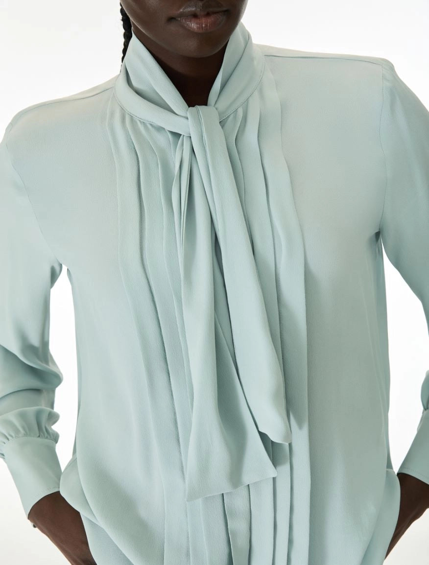 A close-up of a model wearing a pale mint green long-sleeve blouse with a high draped collar and a large front tie, set against a plain white background.