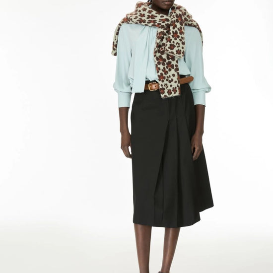 A model stands against a plain white studio background wearing a light blue blouse, a black midi skirt, and a leopard-print scarf draped over her shoulders.