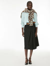 A model stands against a plain white studio background wearing a light blue blouse, a black midi skirt, and a leopard-print scarf draped over her shoulders.
